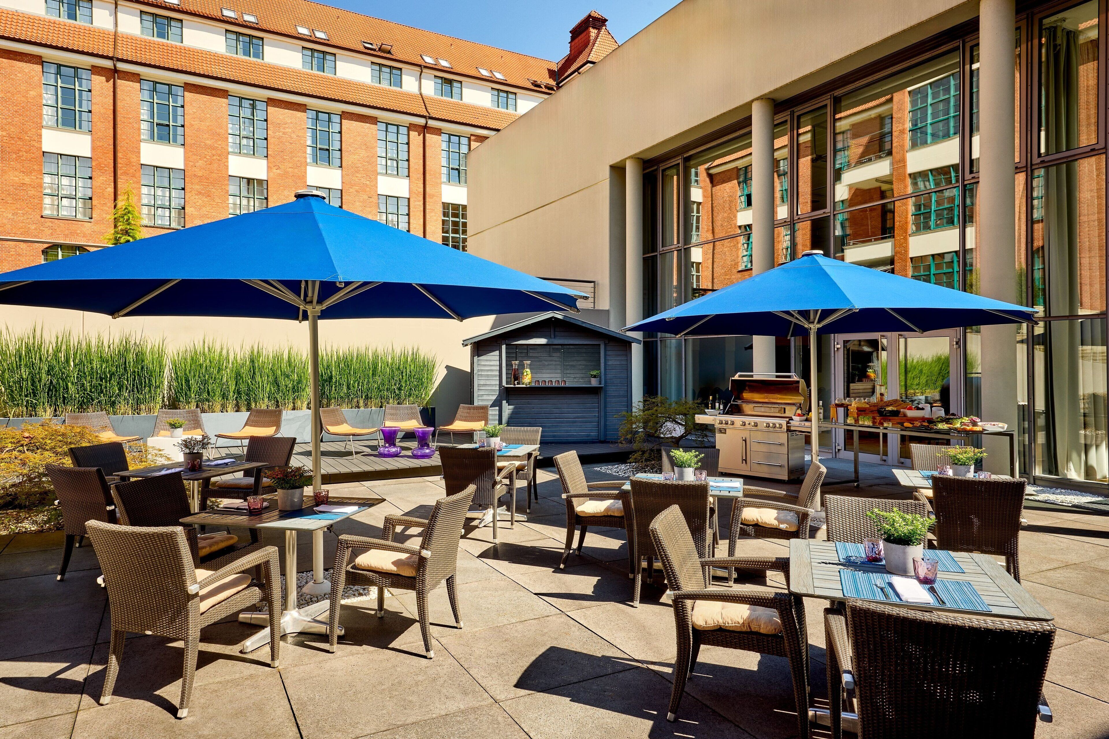 An outdoor patio with wicker furniture and blue umbrellas, a barbecue area, and a building with large windows in the background.