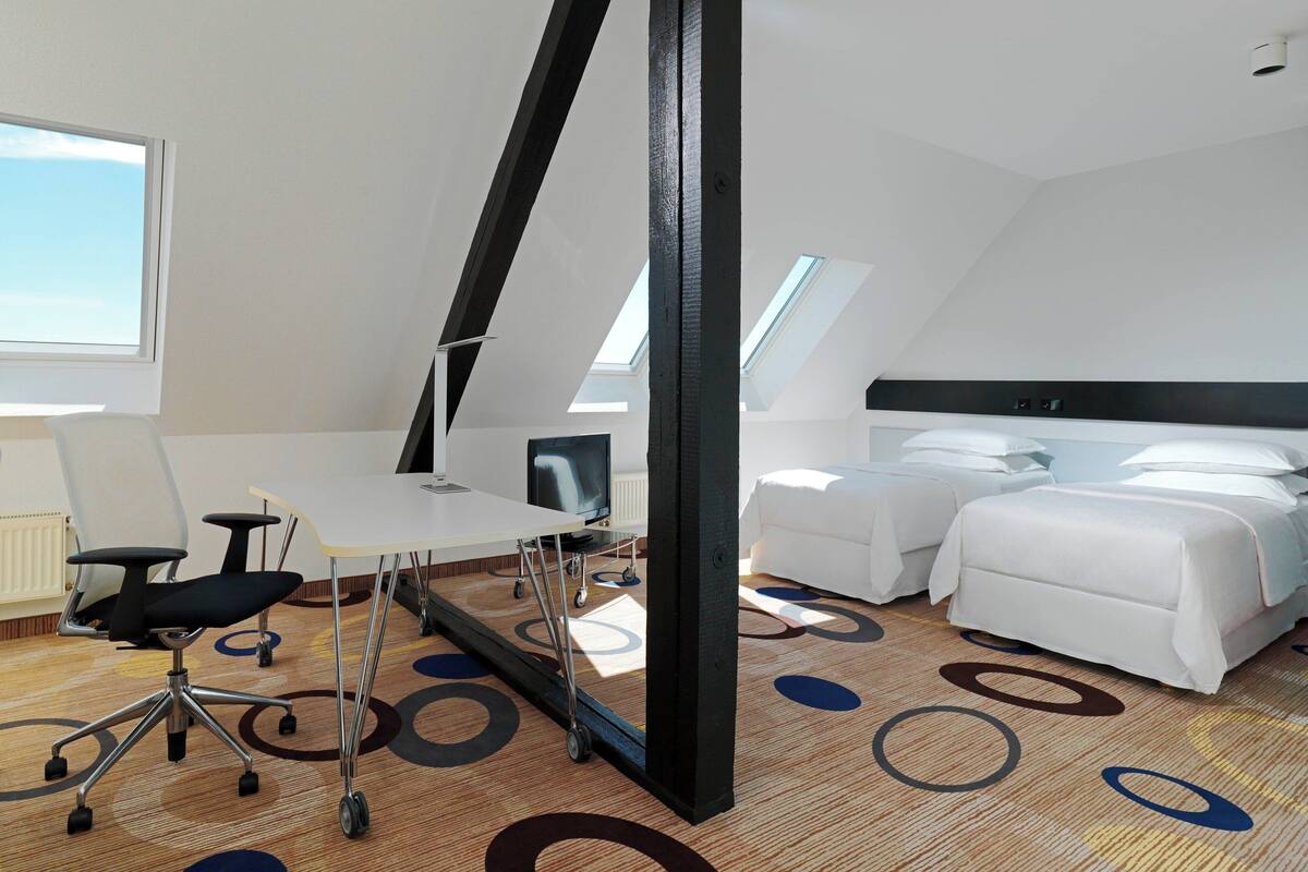 A modern hotel room with a white bed, a desk with a chair, and a large window.