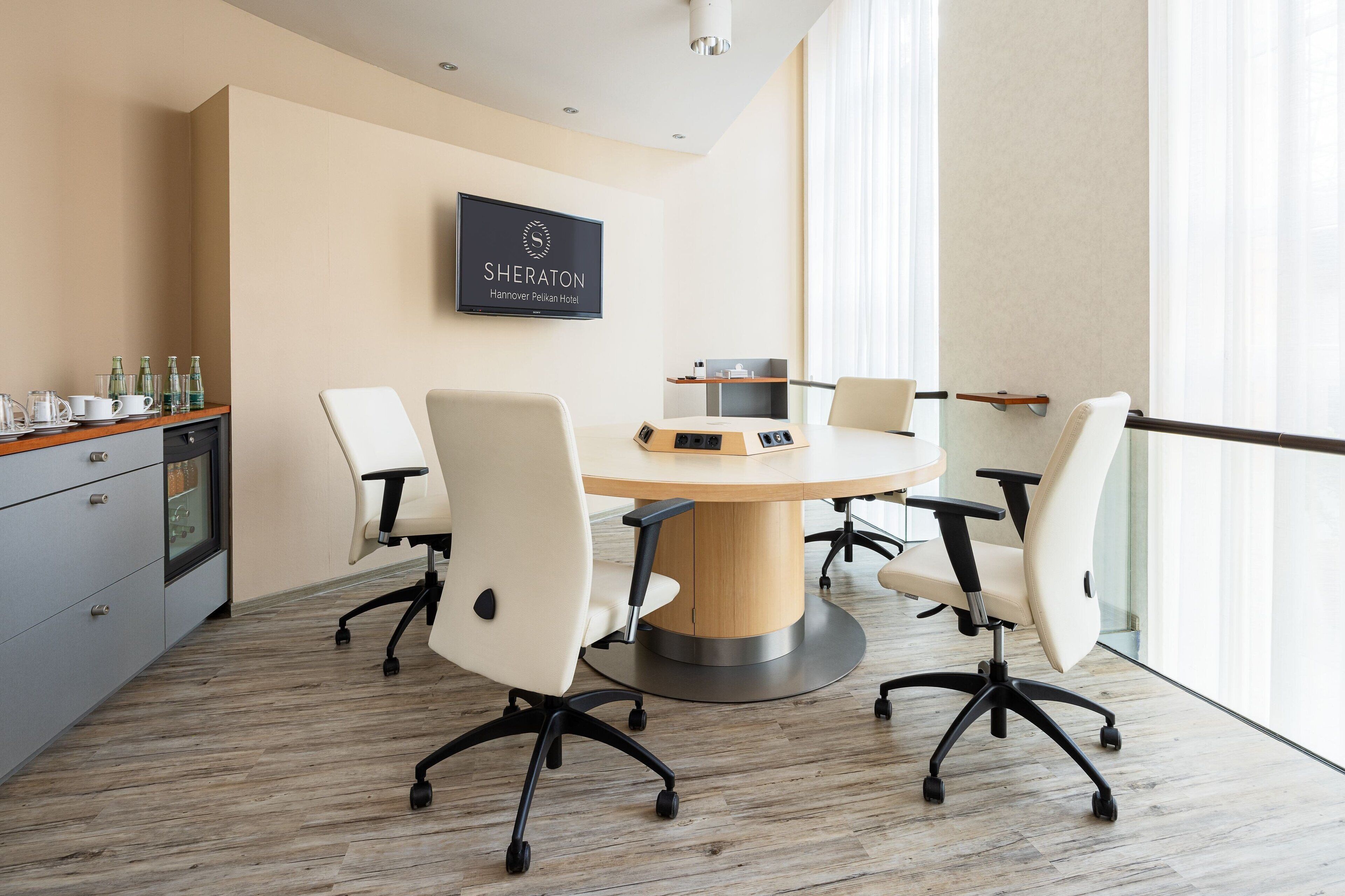 A modern conference room with a round table, four white chairs, a television displaying the Sheraton logo, and a wooden cabinet with glass doors.
