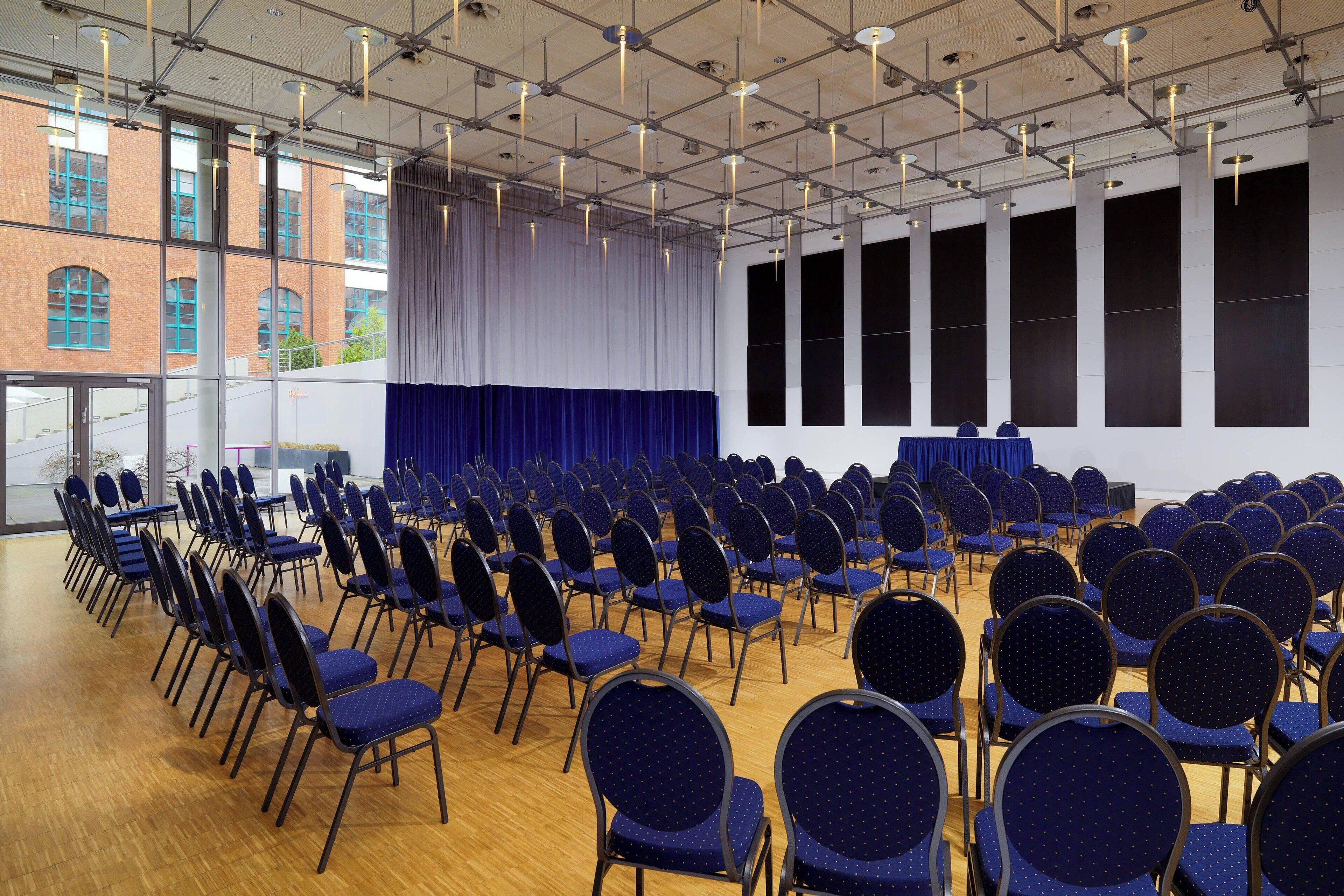 A conference room with rows of chairs, large windows, and a stage with a curtain.