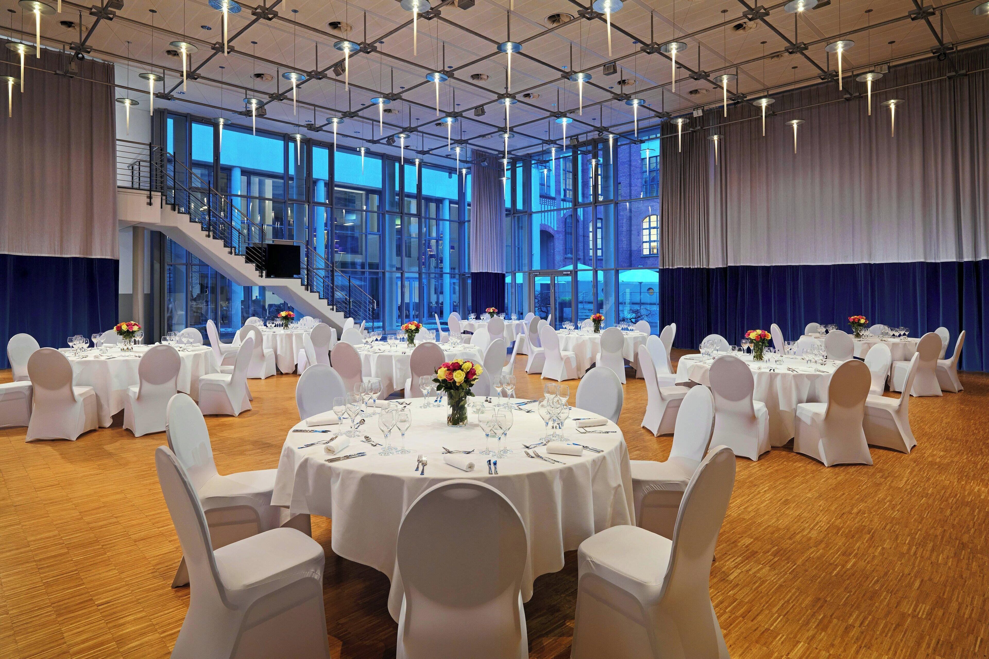 Un salón de banquetes con mesas redondas dispuestas para un evento formal, ventanas amplias y una escalera.