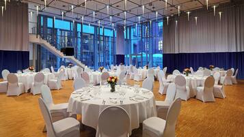 Un salón de banquetes con mesas redondas dispuestas para un evento formal, ventanas amplias y una escalera.