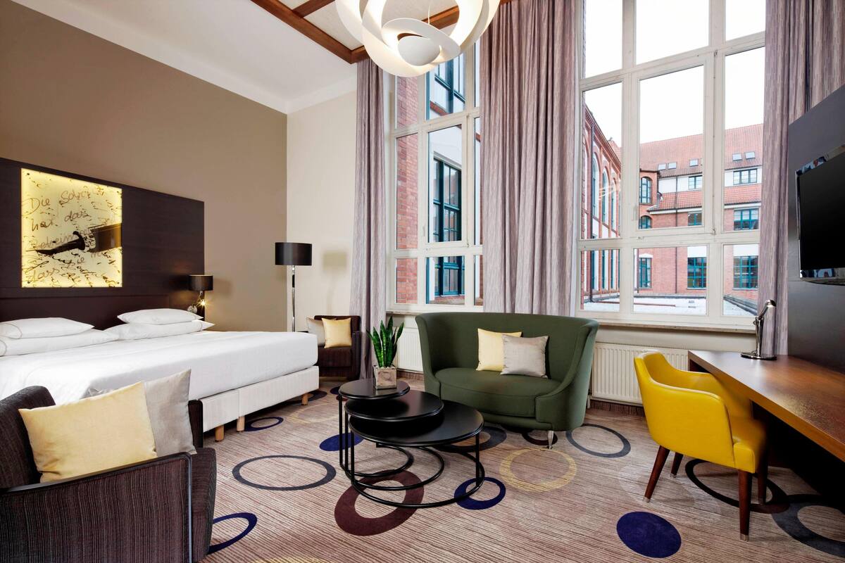 A hotel room with a bed, a green armchair, a yellow chair, a desk, and a large window with a city view.