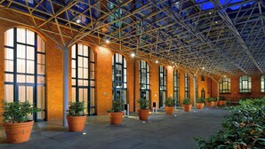 A spacious indoor area with a high ceiling and large windows, featuring brick walls and potted plants.