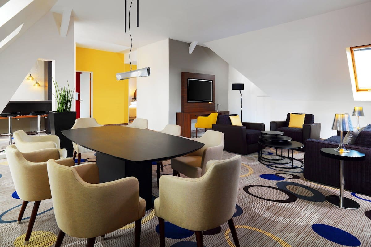 A modern hotel room with a large dining table, comfortable seating, a flat-screen TV, and a cozy seating area with a lamp.