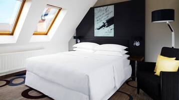 Una habitación de hotel moderna con una cama grande, un escritorio y una silla. Hay tragaluces y un cuadro en la pared.