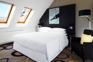 Una habitación de hotel moderna con una cama grande, un escritorio y una silla. Hay tragaluces y un cuadro en la pared.