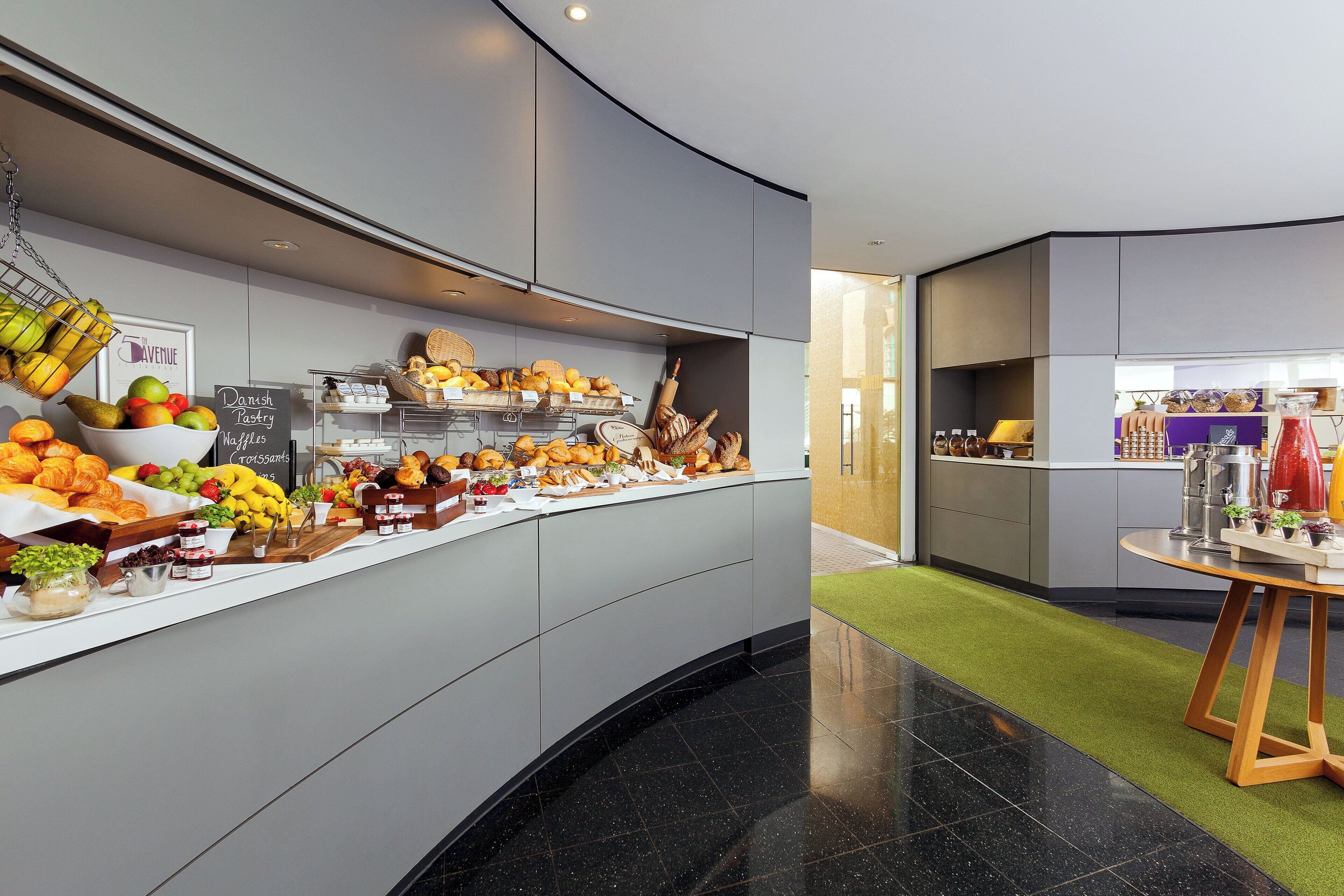 Eine moderne Hotelhalle mit einer gebogenen Rezeption, auf der eine Auswahl an Gebäck, Früchten und Getränken präsentiert wird.