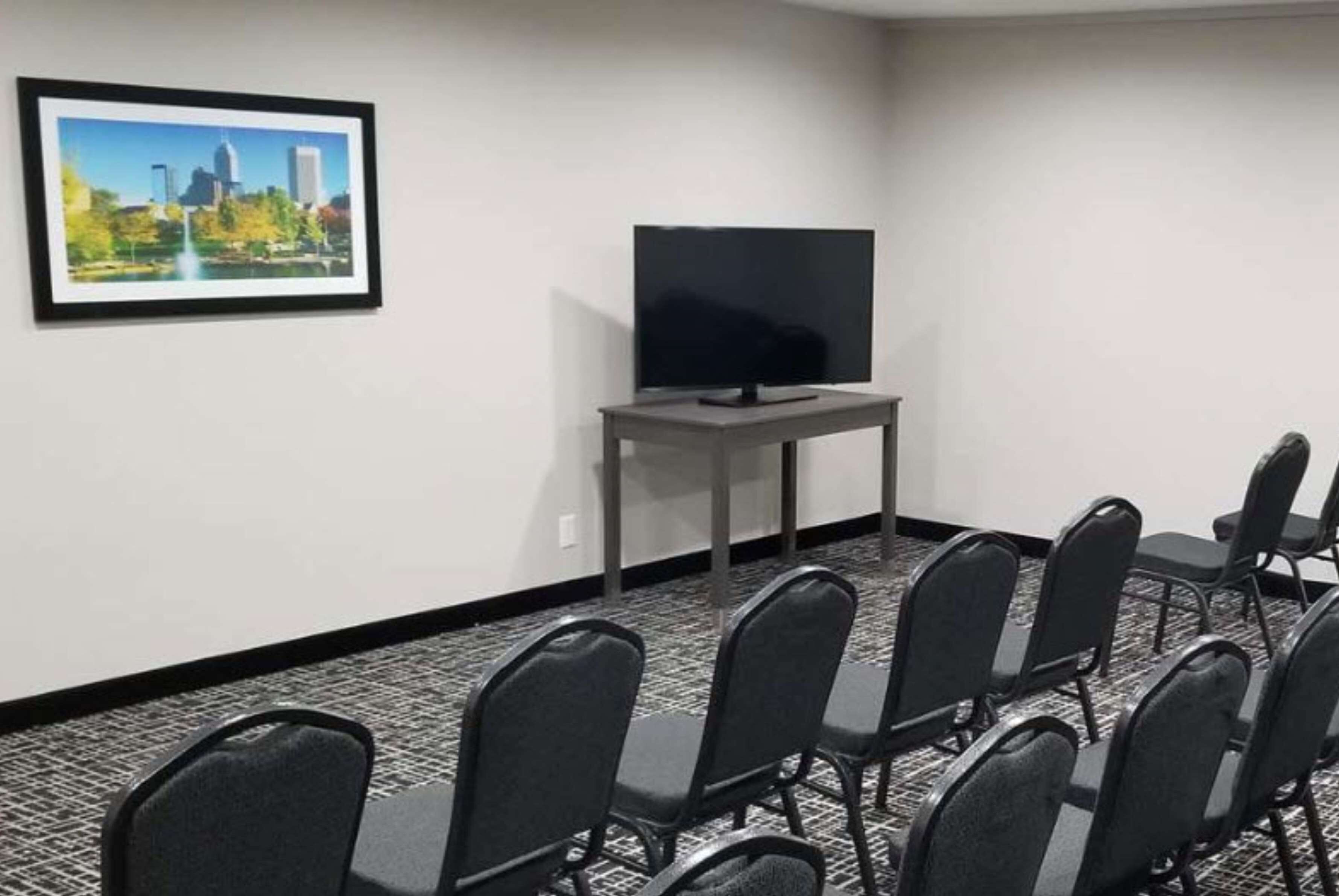 A conference room with a flat-screen TV on a stand, a framed cityscape picture on the wall, and rows of black chairs.