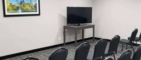 A conference room with a flat-screen TV on a stand, a framed cityscape picture on the wall, and rows of black chairs.