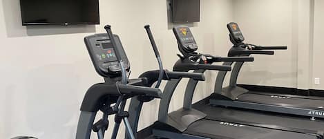 Three exercise machines with digital displays, a wall-mounted TV, and a plain wall.