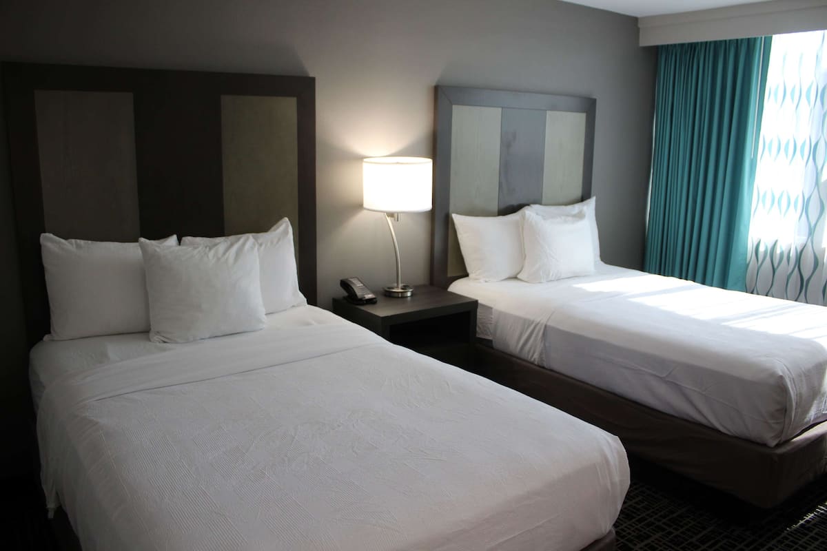 A hotel room with two single beds, a nightstand with a lamp, and a window with curtains.