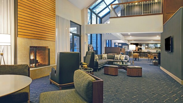 A spacious lobby with a fireplace, comfortable seating, a staircase to an upper level, and a view of the city through large windows. - Courtyard Chicago Elmhurst/Oakbrook Area (Elmhurst)