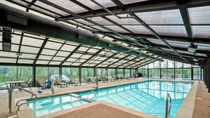An indoor swimming pool with clear glass walls, metal beams, and lounge chairs. - Courtyard Chicago Elmhurst/Oakbrook Area (Elmhurst)