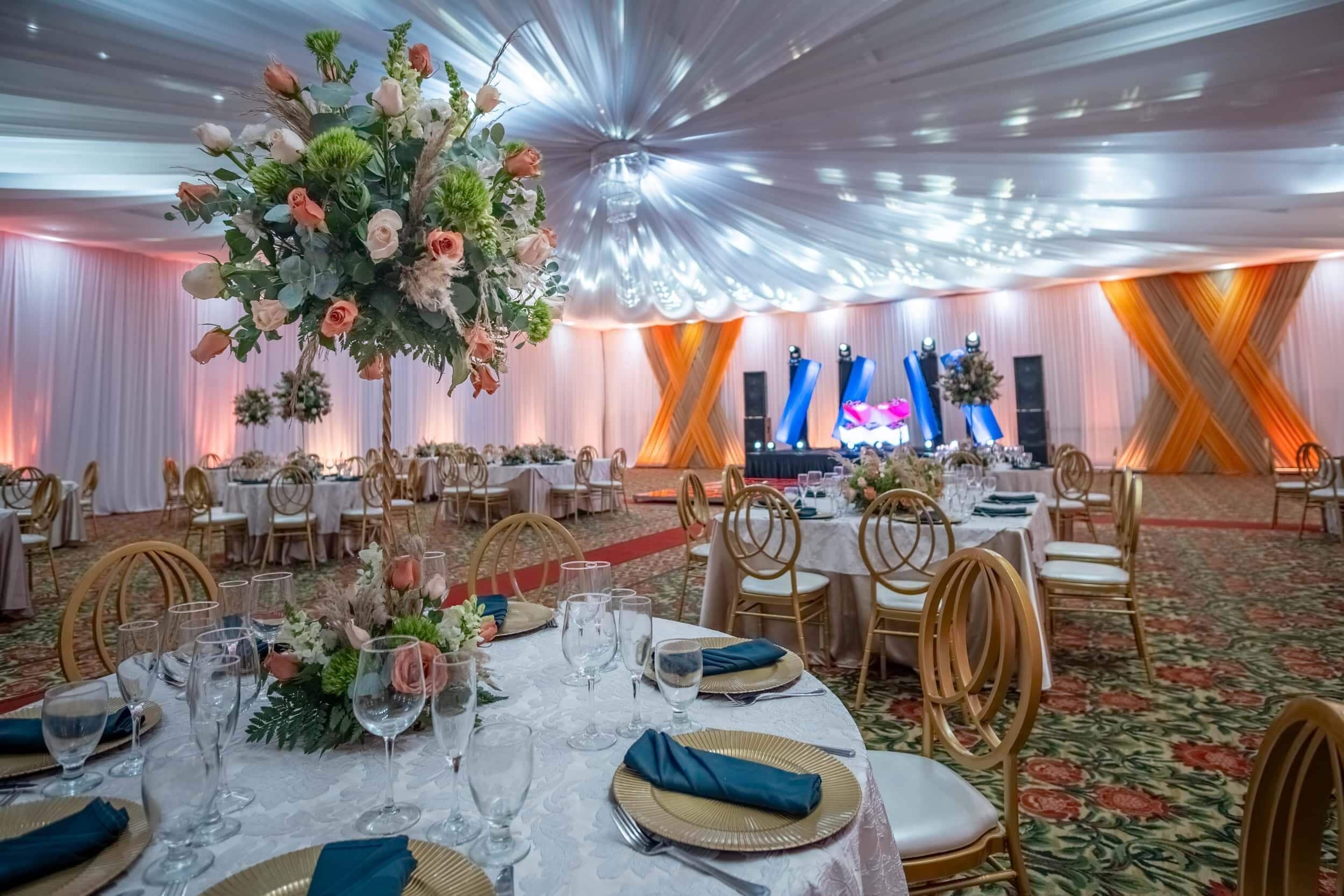 A banquet hall with round tables set for a formal event, featuring floral centerpieces and gold-rimmed glassware.