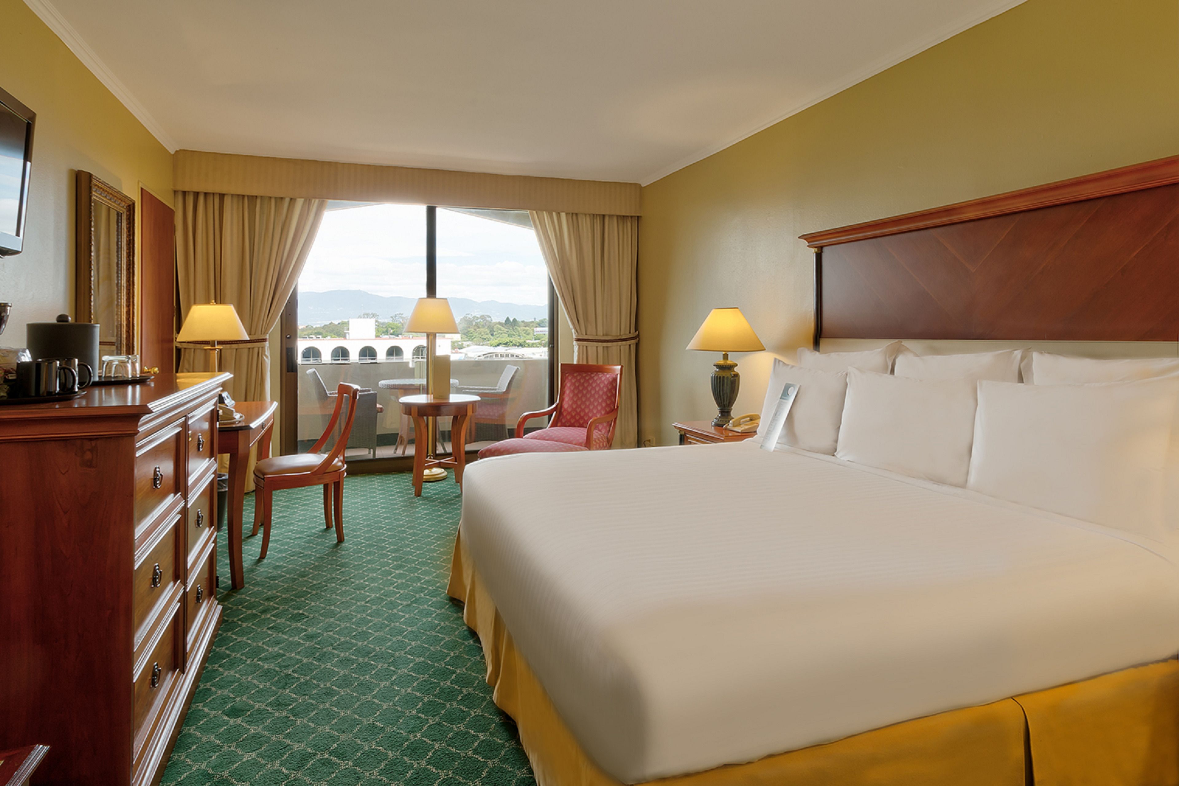 A hotel room with a large bed, a desk with a lamp, a chair, and a view of the city through large windows.