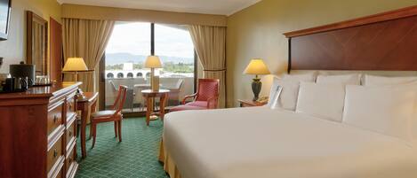 Habitación de hotel con una cama grande, un escritorio con lámpara, una silla y vistas a la ciudad a través de amplias ventanas.