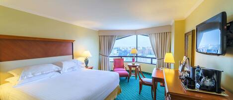 Quarto de hotel com uma cama grande, uma mesa com cafeteira, uma cadeira e uma televisão.