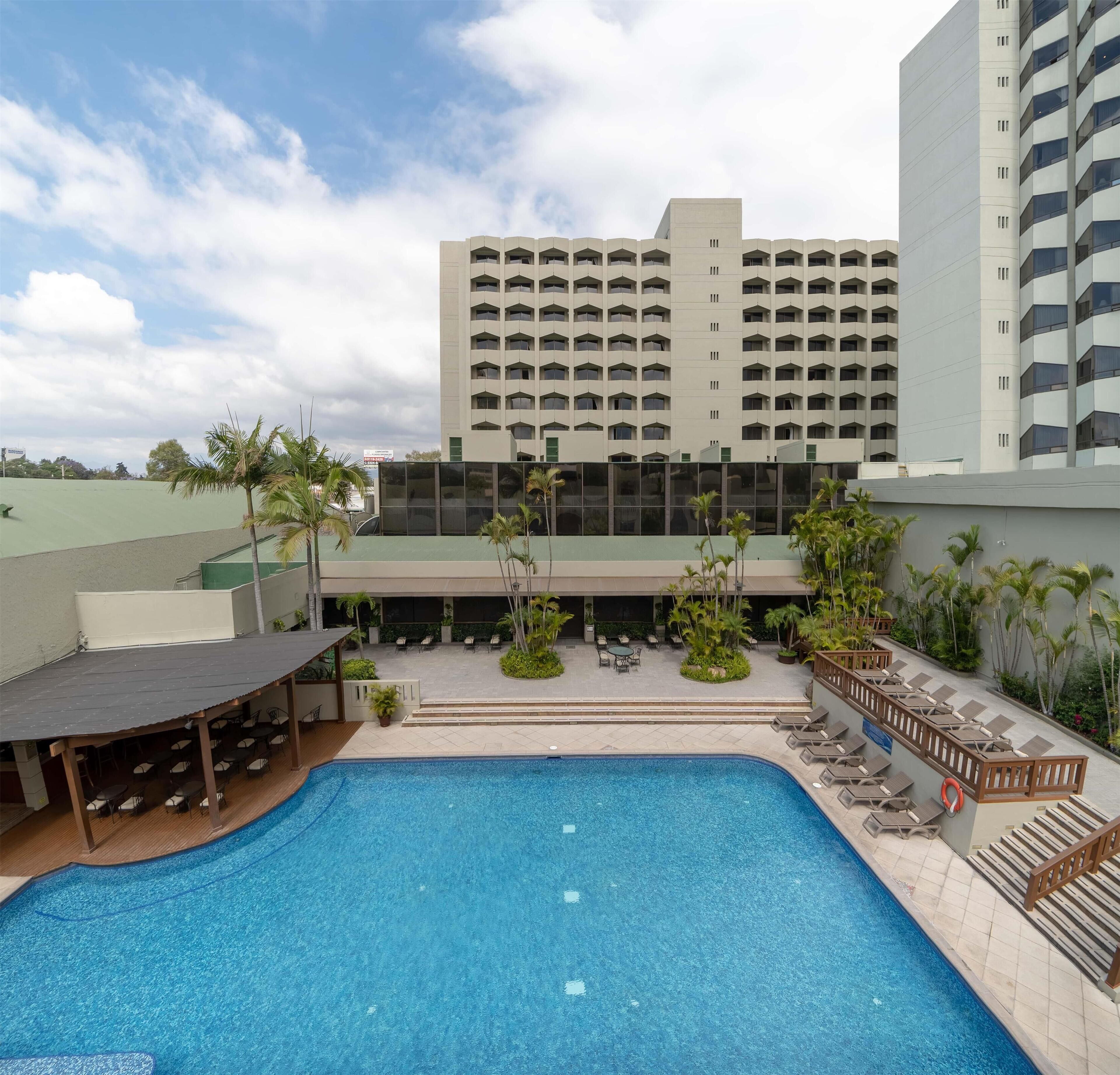 A swimming pool with lounge chairs and a covered seating area, surrounded by palm trees and a large hotel building in the background.