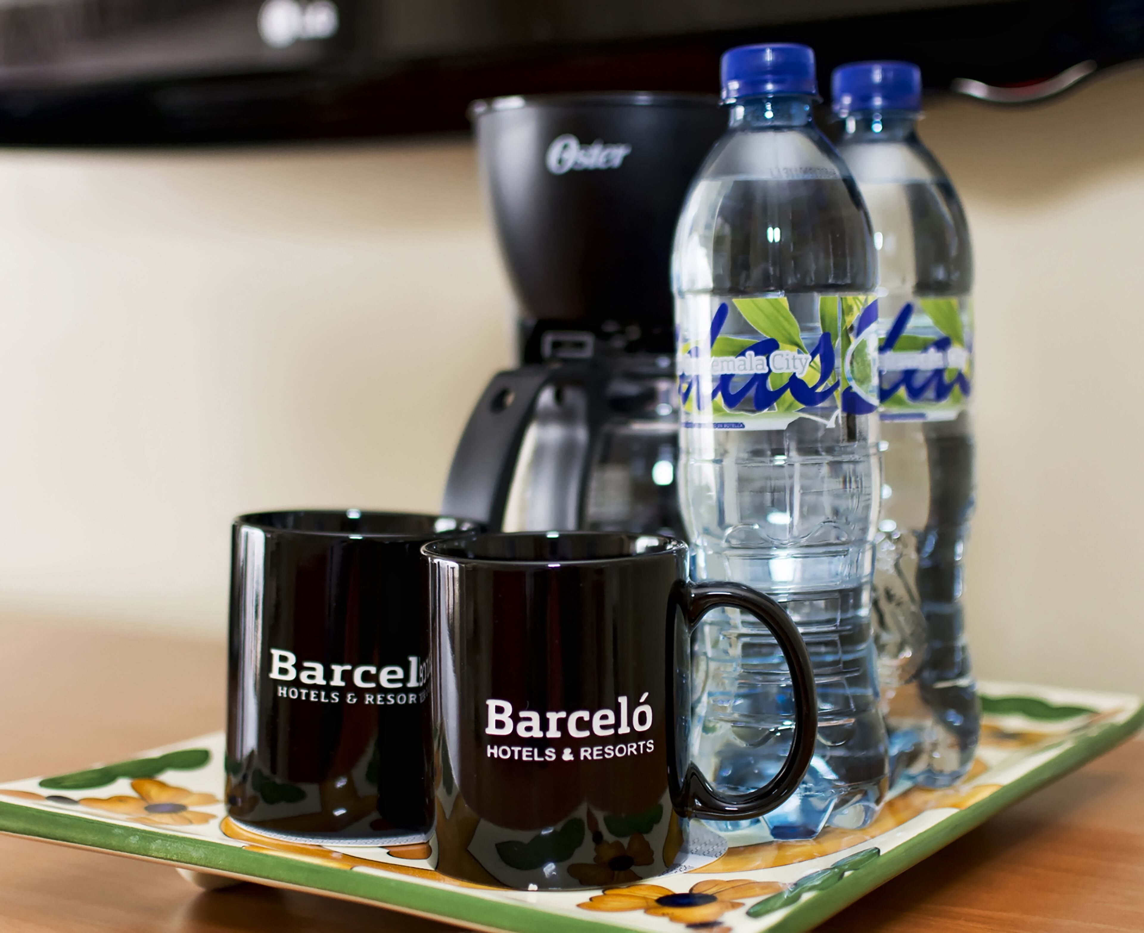 Un tavolo con due tazze nere con la scritta "Barceló Hotels & Resorts", una macchina del caffè e due bottiglie d'acqua con etichetta blu.