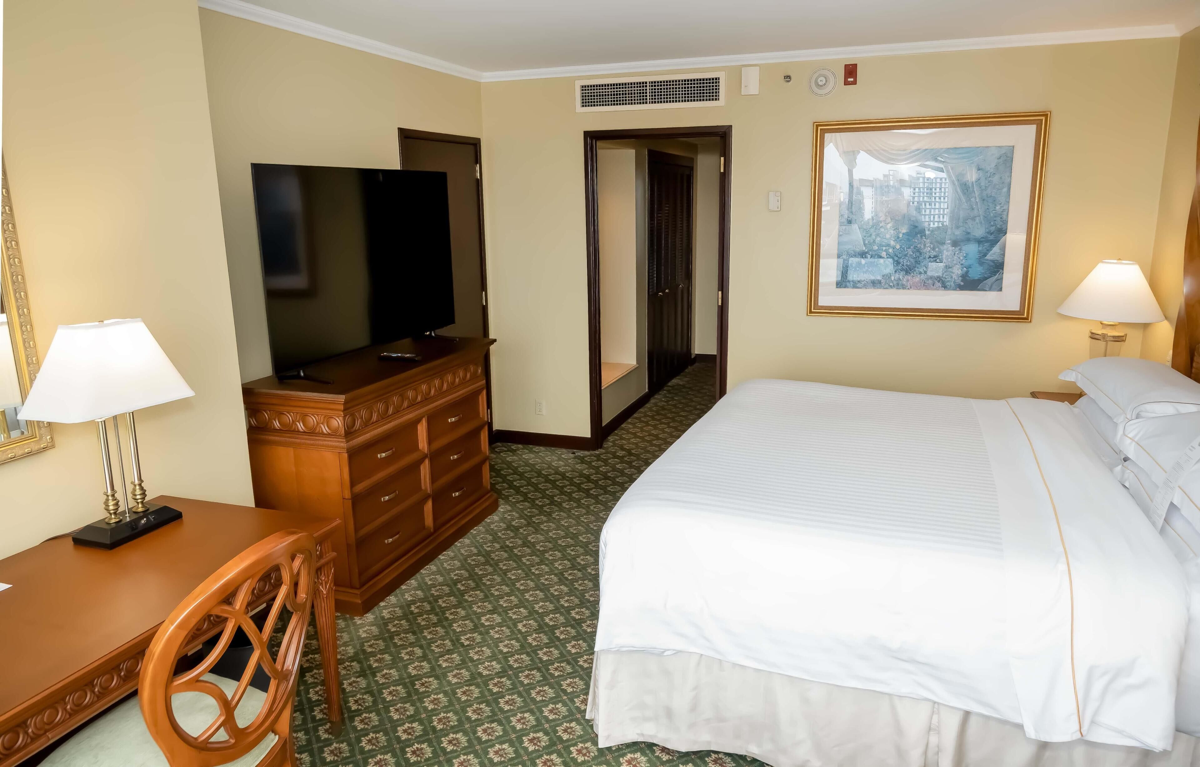 A hotel room with a bed, a desk with a lamp, a dresser, a television, and a painting on the wall.