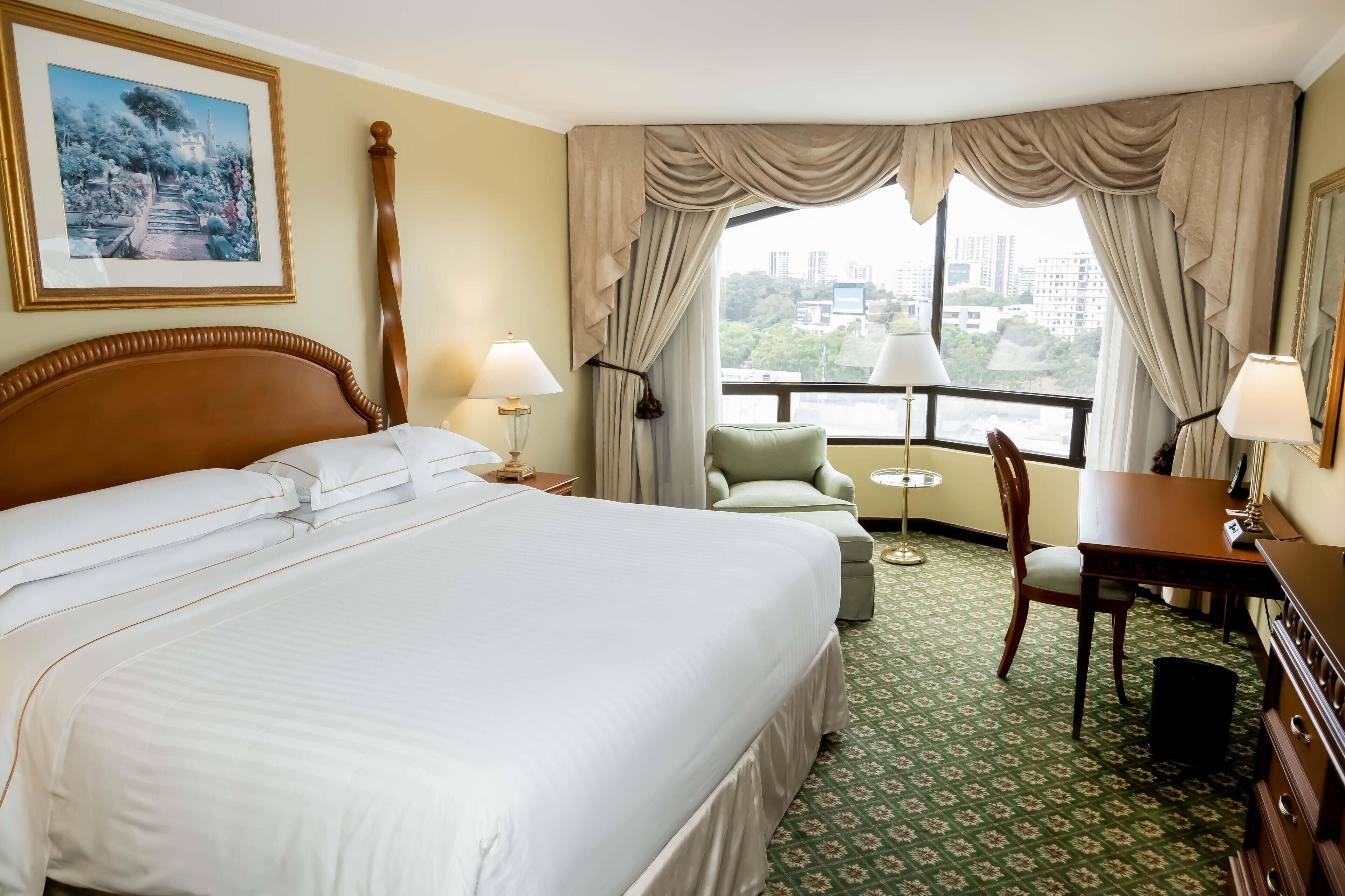 Habitación de hotel con una cama grande, dos mesitas de noche, una silla, una pequeña mesa redonda y vistas a la ciudad a través de las ventanas.