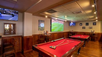 A pool table with a red felt surface, a billiard cue, and a set of balls. There are two wooden chairs and a small table with a drink menu.