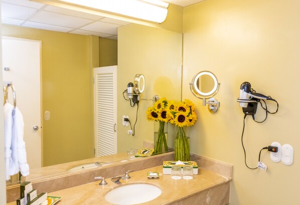 A bathroom with a sink, mirror, and two hair dryers mounted on the wall. Sunflowers are placed on the counter.