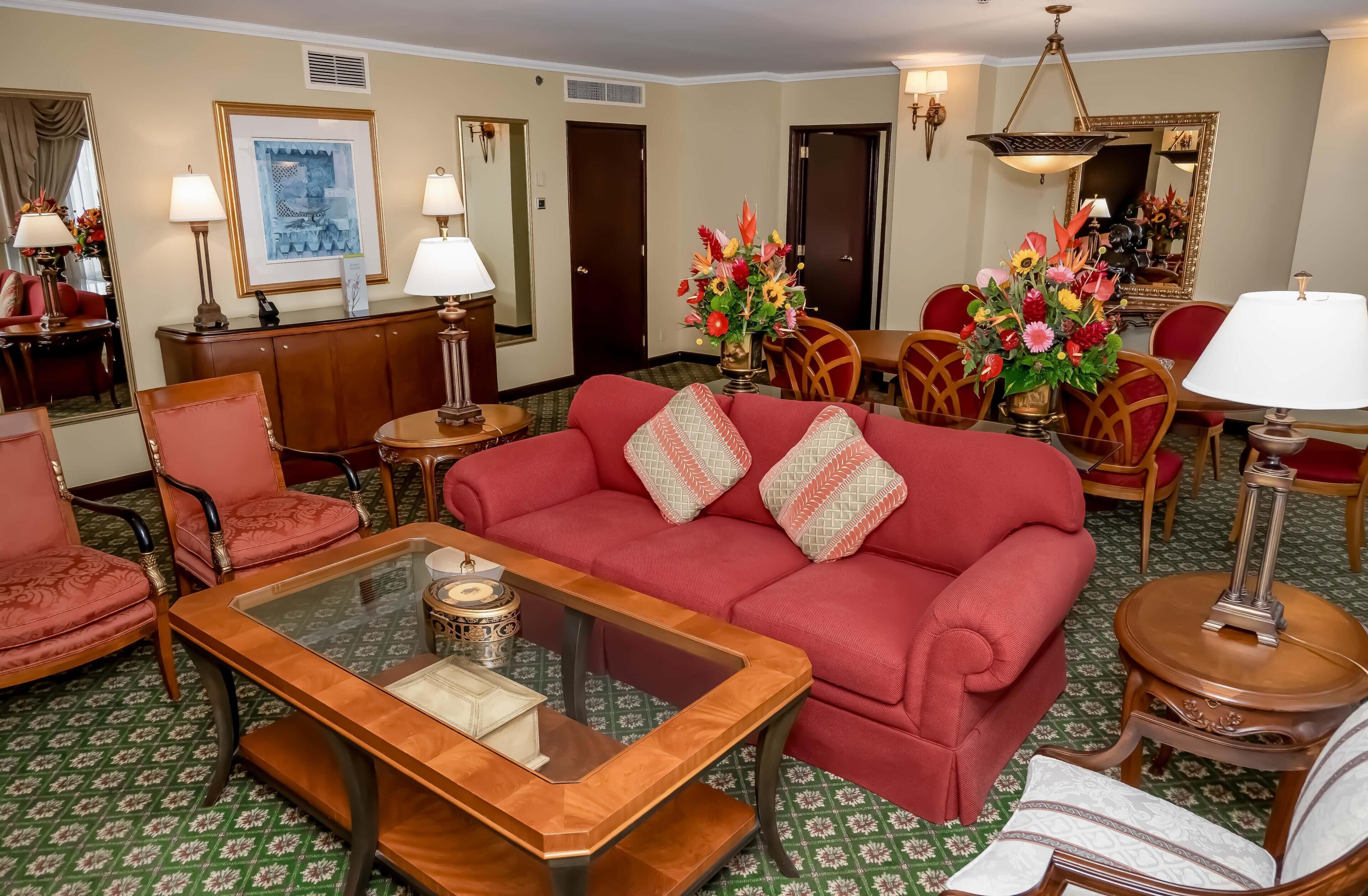 A hotel room with a red sofa, a glass coffee table, and a dining area with a table and chairs.