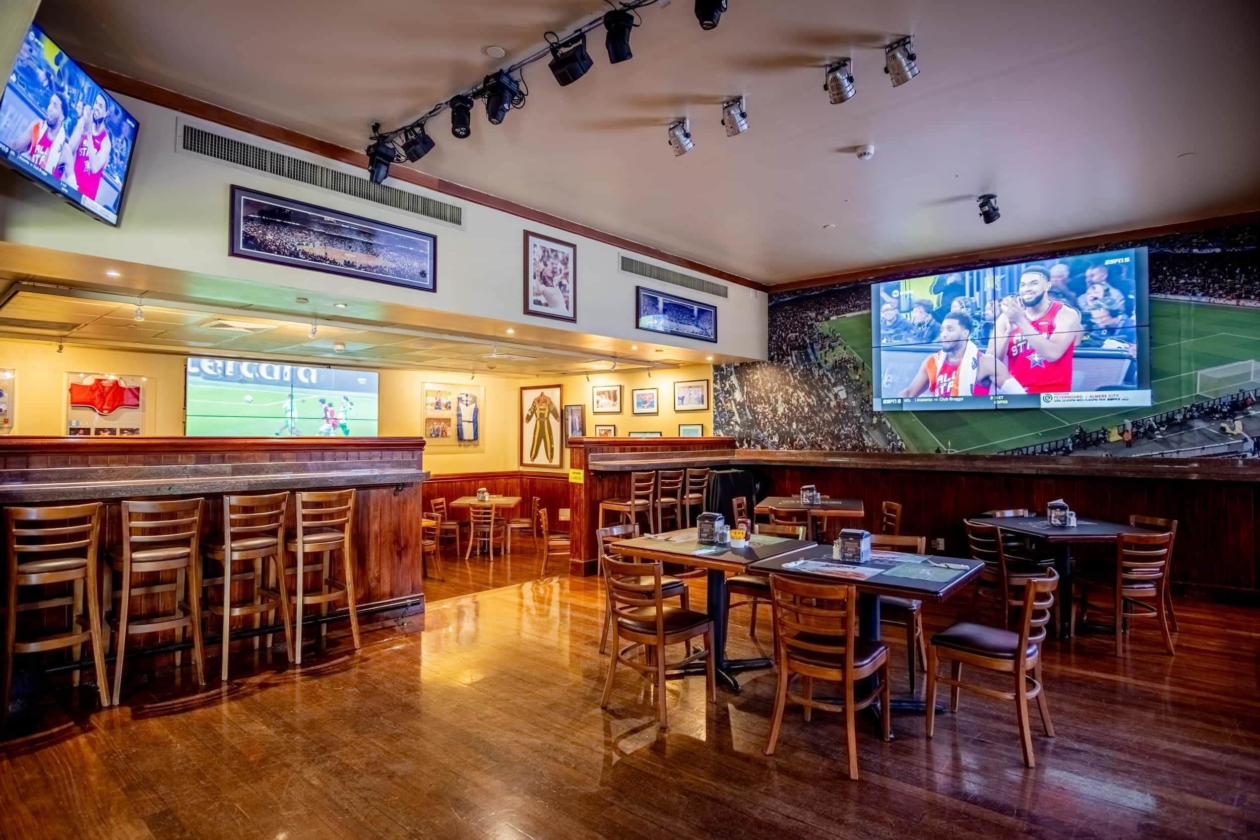 Un espace bar avec des meubles en bois, un grand écran diffusant des événements sportifs et des tableaux encadrés aux murs.