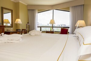 Ein Hotelzimmer mit einem großen Bett, zwei Nachttischen, einem Schreibtisch mit Stuhl, einem Spiegel und einem Fenster mit Stadtblick.