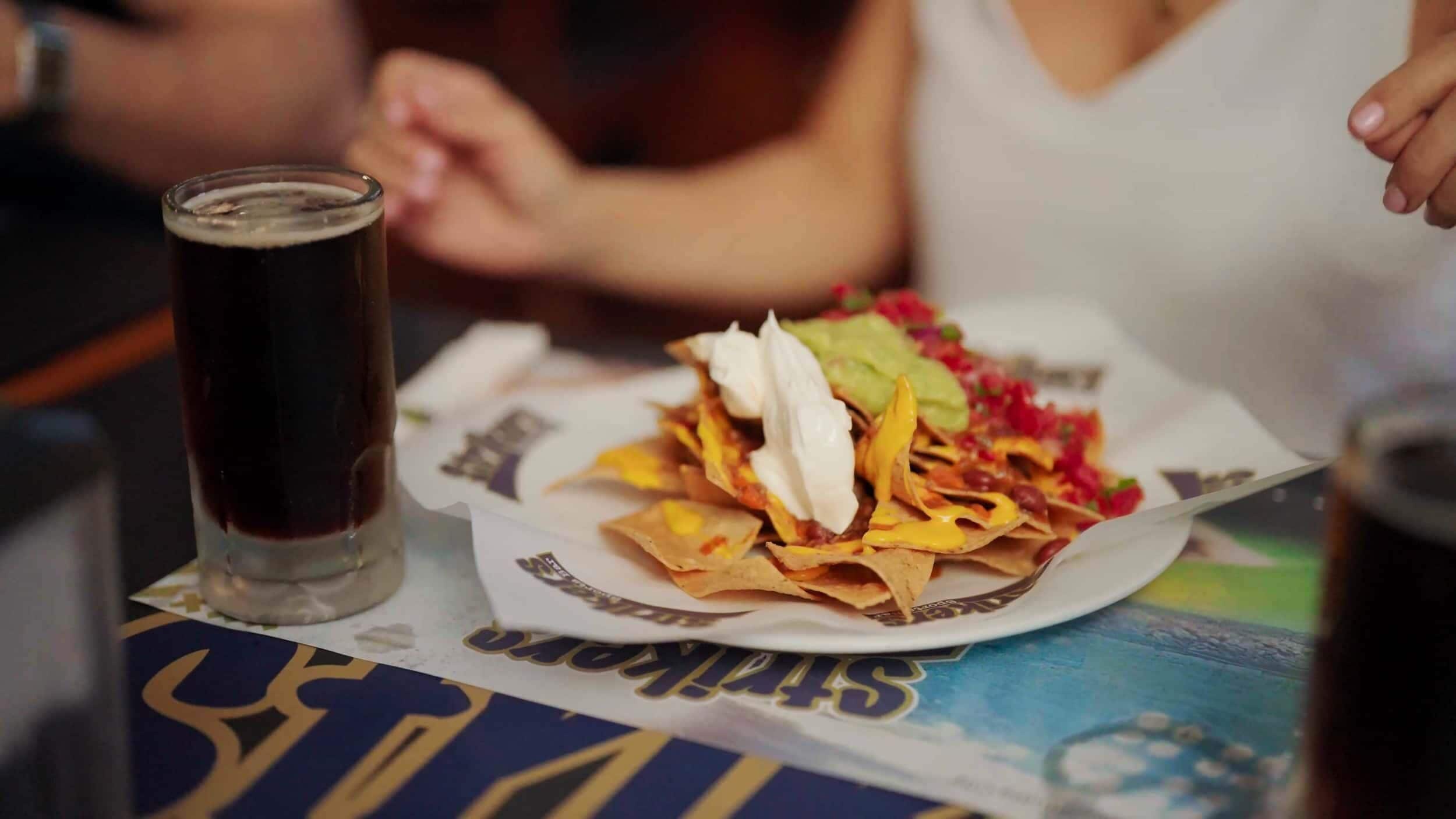 A plate of nachos with toppings and a glass of dark beverage on a table.