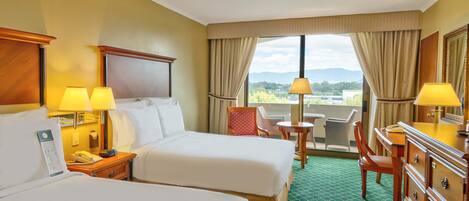 A hotel room with two beds, a desk with a lamp, a chair, a dresser, and a view of the outdoors.