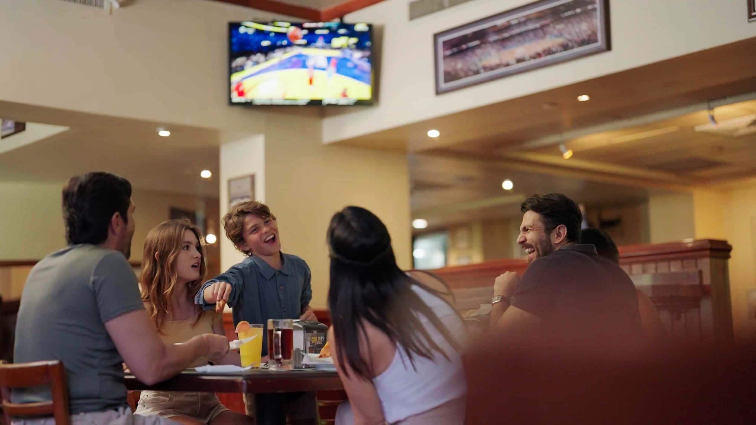 A group of people dining in a restaurant with a television mounted on the wall displaying a sports event.
