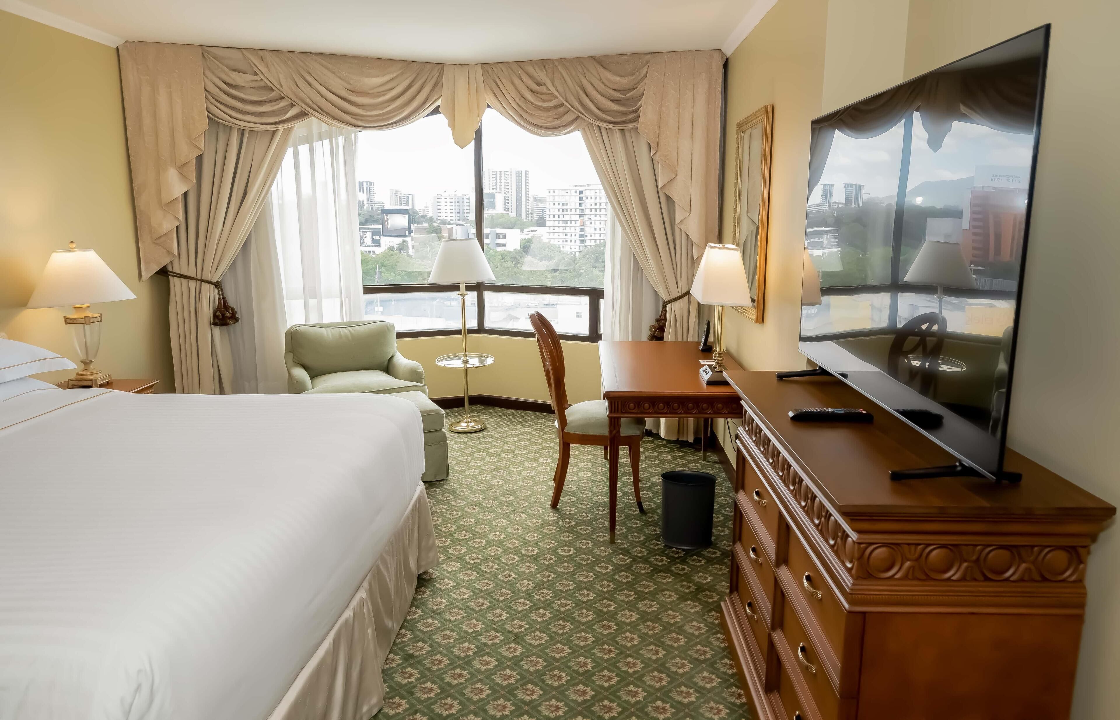 Habitación de hotel con cama, escritorio, silla, televisión y vistas de la ciudad a través de amplias ventanas.
