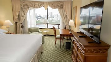 Habitación de hotel con cama, escritorio, silla, televisión y vistas de la ciudad a través de amplias ventanas.