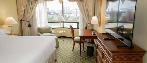 Habitación de hotel con cama, escritorio, silla, televisión y vistas de la ciudad a través de amplias ventanas.