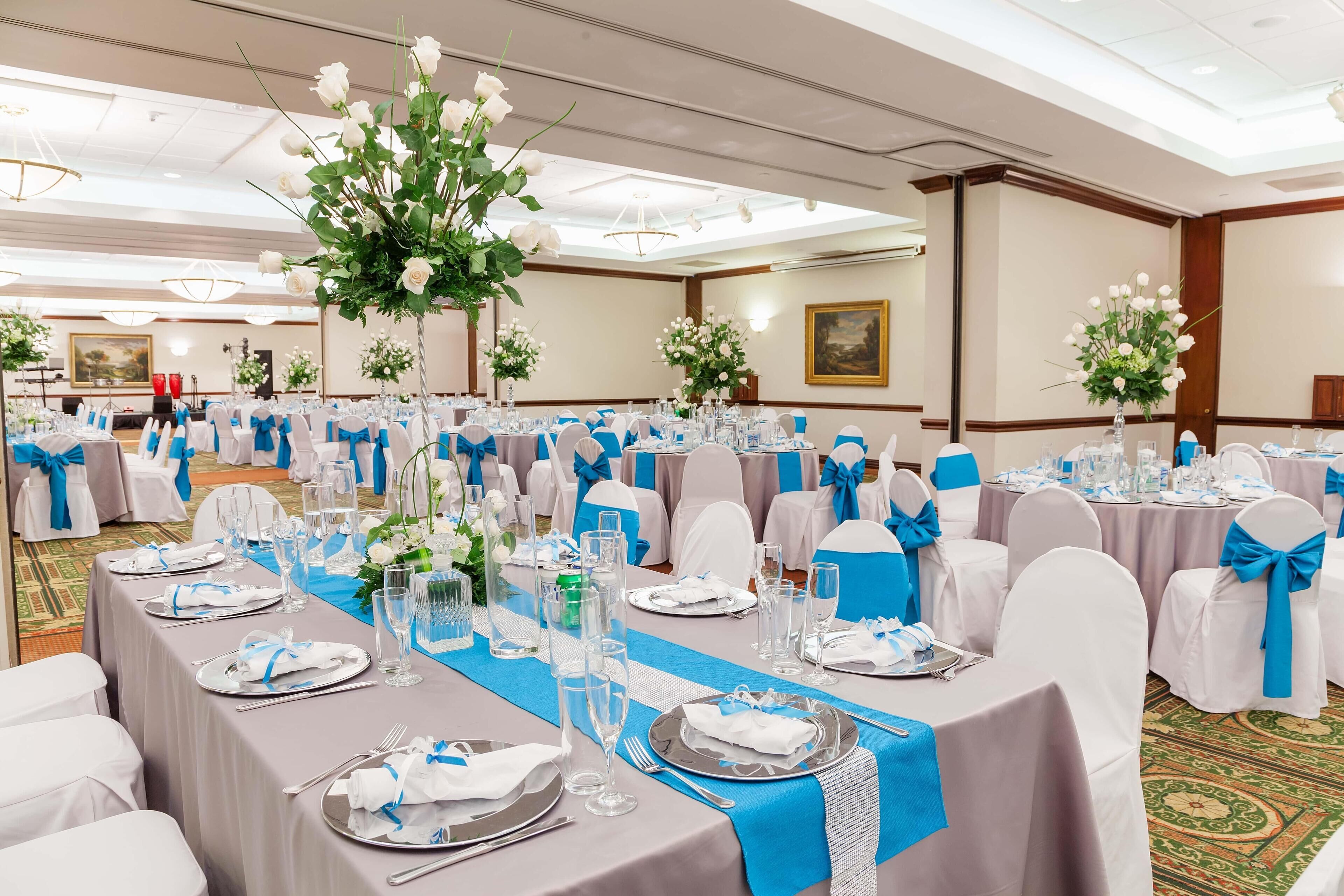 Une salle de banquet où les tables sont dressées pour un événement formel, avec une décoration aux tons de blanc et de bleu, des arrangements floraux en guise de centre de table et un sol recouvert de moquette.
