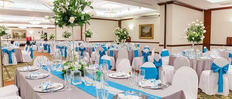 Salão de banquetes com mesas montadas para um evento formal, decoração em branco e azul, centros de mesa florais e piso coberto de carpete.