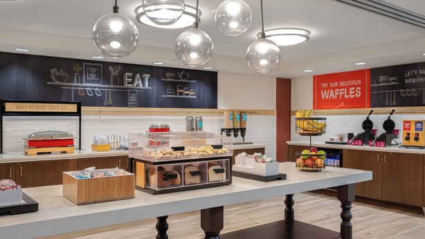 A breakfast buffet area with clear glass containers of food, a sign promoting waffles, and a wall with kitchen utensils illustrations. - Hampton Inn Dubuque (Dubuque)