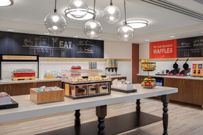 A breakfast buffet area with clear glass containers of food, a sign promoting waffles, and a wall with kitchen utensils illustrations.