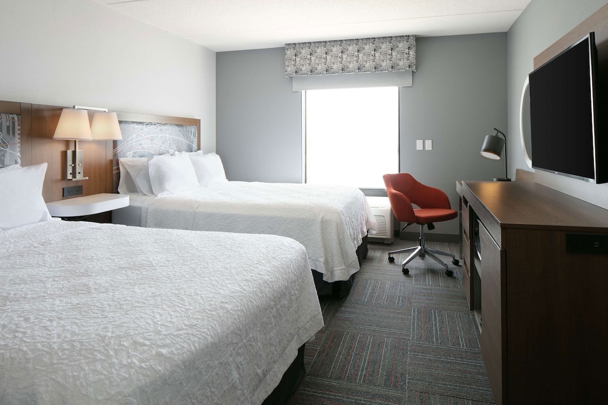 A hotel room with two beds, a desk, a chair, a television, and a large window.