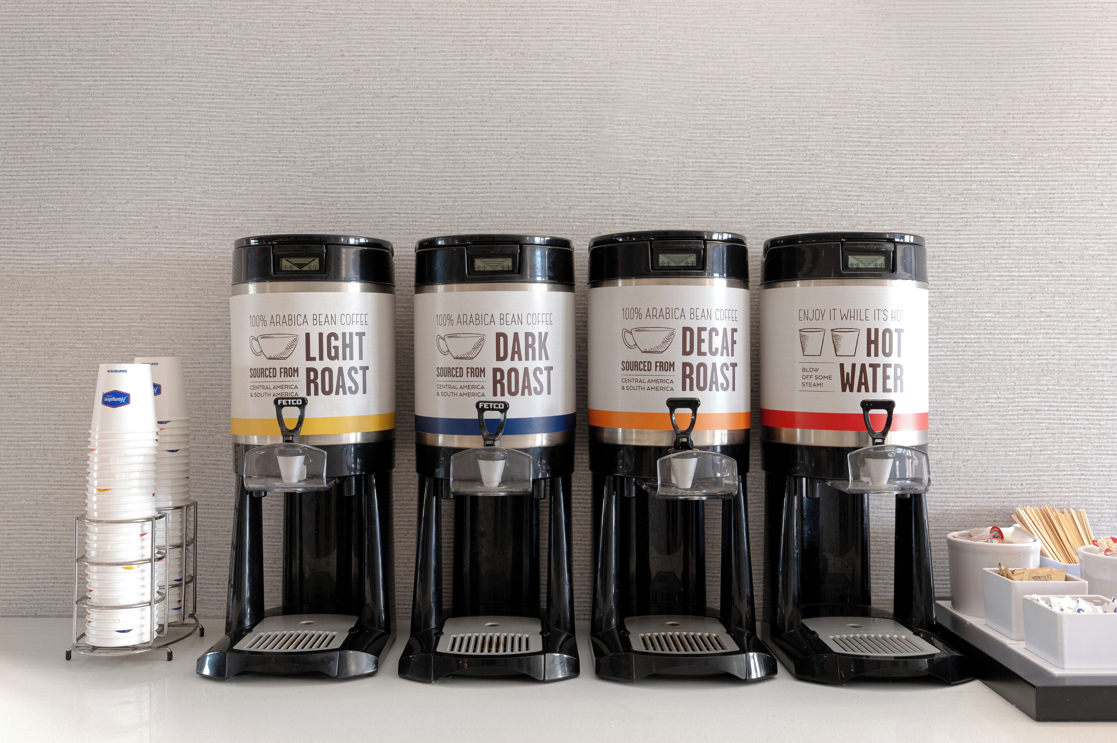 Quatre distributeurs de café, chacun portant une étiquette indiquant le type de torréfaction, et une pile de gobelets jetables.