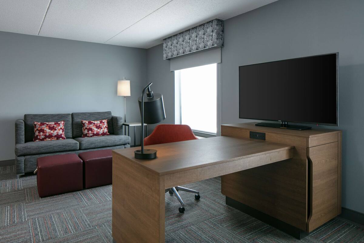 A modern hotel room with a grey sofa, red ottomans, a wooden desk, a flat-screen TV, and a lamp.