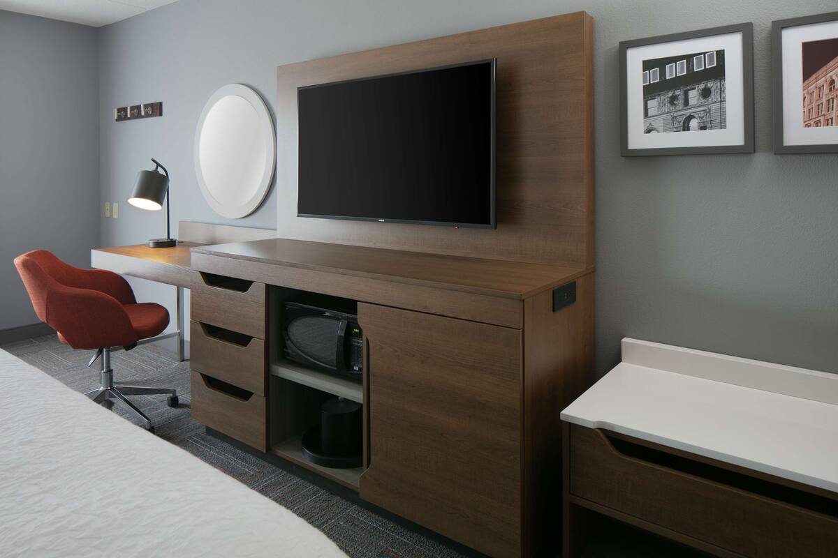 A modern hotel room with a wooden desk, a flat-screen TV, a red chair, and a bed.