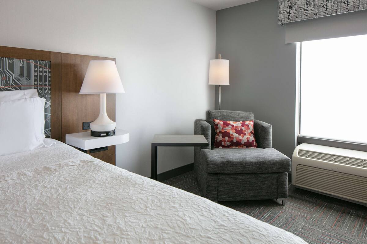 A hotel room with a bed, a nightstand with a lamp, a small table, a gray armchair with a patterned pillow, and a window with an air conditioning unit.
