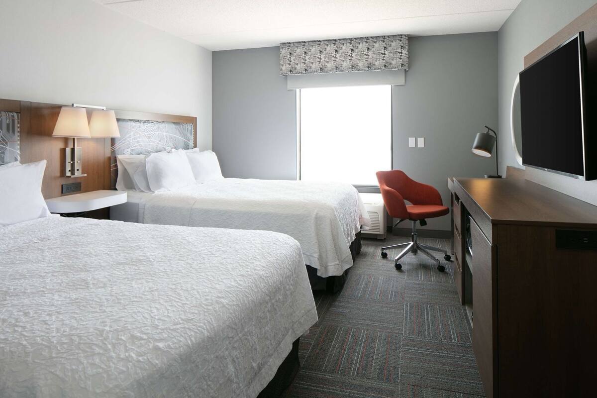 A hotel room with two beds, a desk, a chair, a television, and a large window.