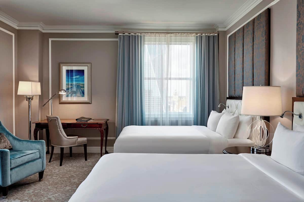 A hotel room with two beds, a desk, a chair, and a lamp.