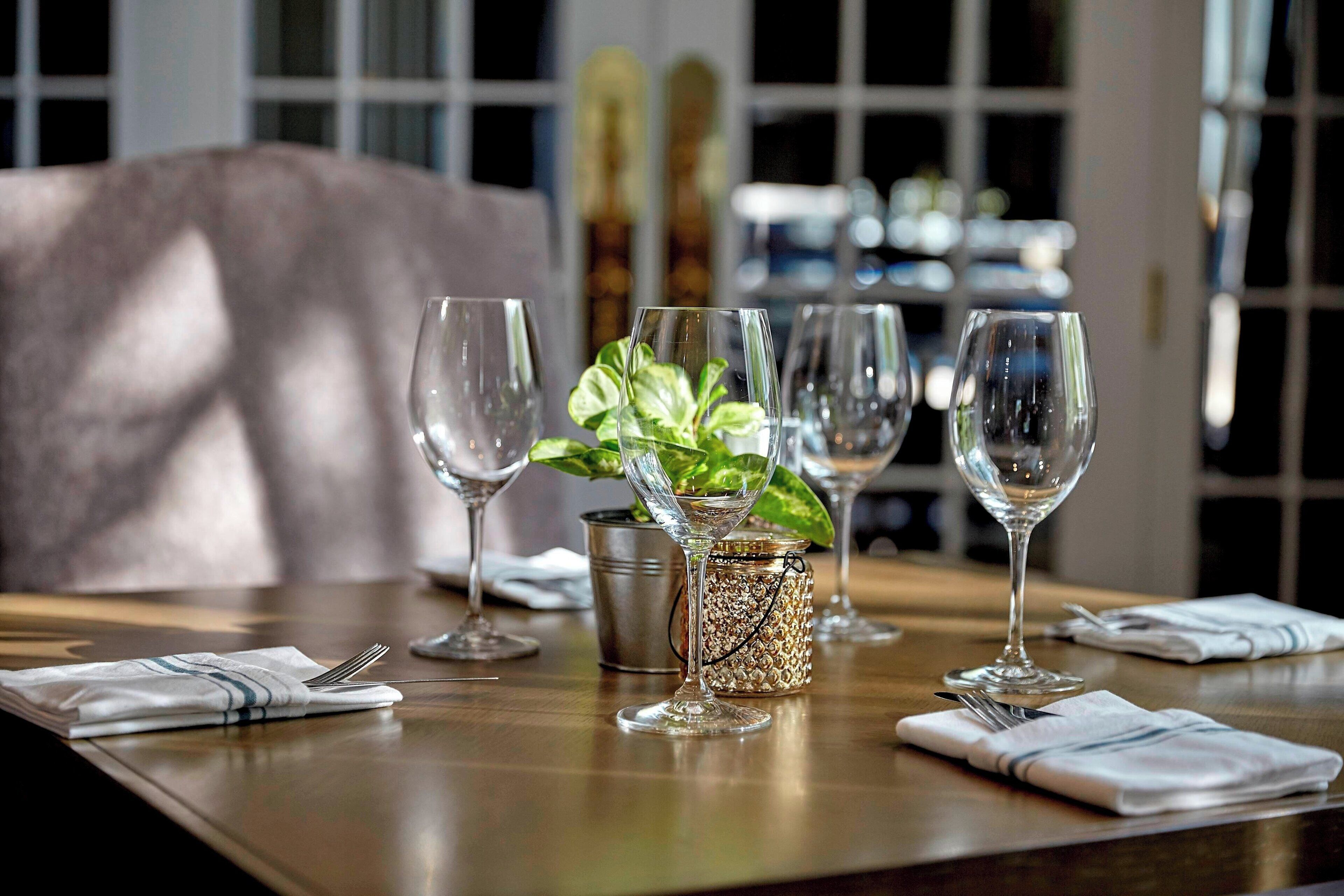 Una mesa de comedor dispuesta con copas de vino, una planta y un recipiente decorativo.