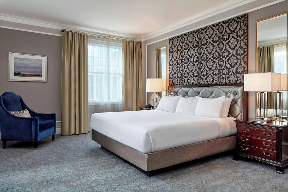 A hotel room with a large bed, a blue armchair, a nightstand with lamps, and a patterned wallpaper.